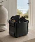 Panier de rangement en cuir noir utilisé comme vide-poche sur un meuble en bois, avec montre et lunettes face à la mer