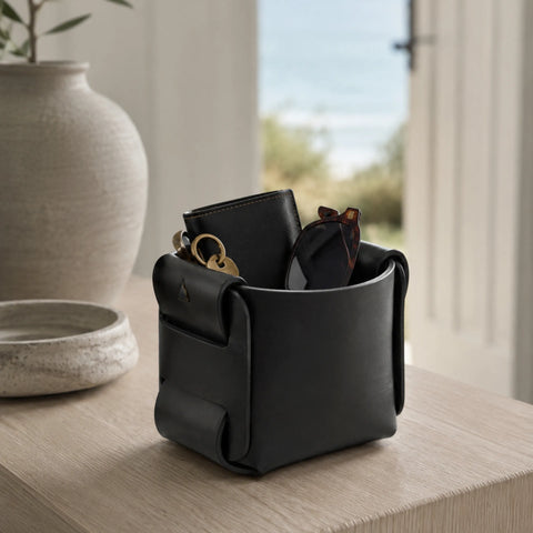 Panier de rangement en cuir noir utilisé comme vide-poche sur un meuble en bois, avec montre et lunettes face à la mer